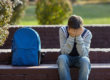 teen boy with hands over face sitting next to backpack showing signs of drug use