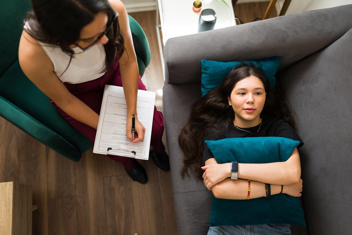 a teen girl lays down on a couch holding a pillow and talking to a therapist in clinical therapy